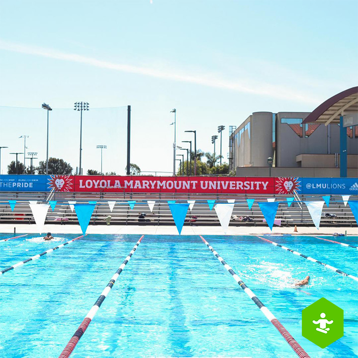 Outdoor view of Loyola Marymount University’s swimming pool, with multiple lap lanes stretching across the foreground. Blue and white lane markers and triangular pennant flags hang above the water, while a large banner displaying Loyola Marymount University spans the bleachers in the background under a clear sky.