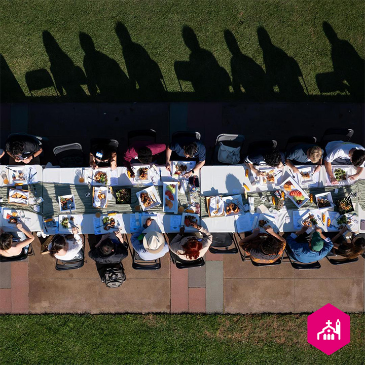 Aerial view of a long outdoor community meal where attendees are seated on both sides of a banquet table filled with plates of food and drinks. Long shadows stretch across the adjacent lawn in the late afternoon sun, highlighting gathering, reflection, and con