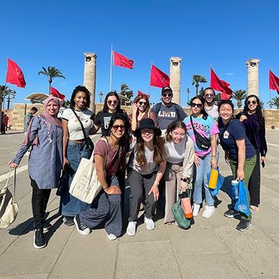 Students and staff pose outside in front of red flags and a blue sky on an AB trip to Morocco.
