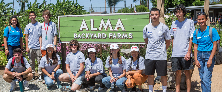 A group of students in grey t-shirts gather in front of a green sign that says Alma Backyard Farms outside.