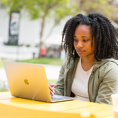 A student sits outside at a yellow table looking at a laptop.