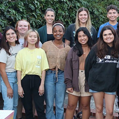 A group of students gathers outside in front of green bushes in the backyard of the service house.