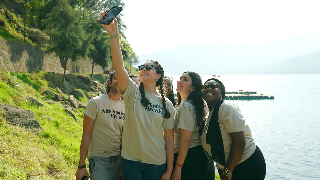 A group of students pose with a phone for a selfie with a green tree and blue sky in the background.