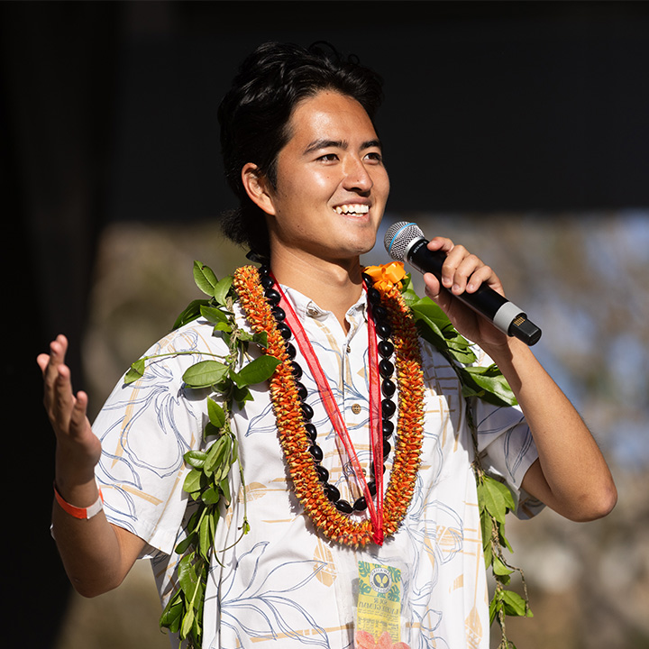 A male student in a Hawaiian shirt and lei stands outside on a stage holding a mic.