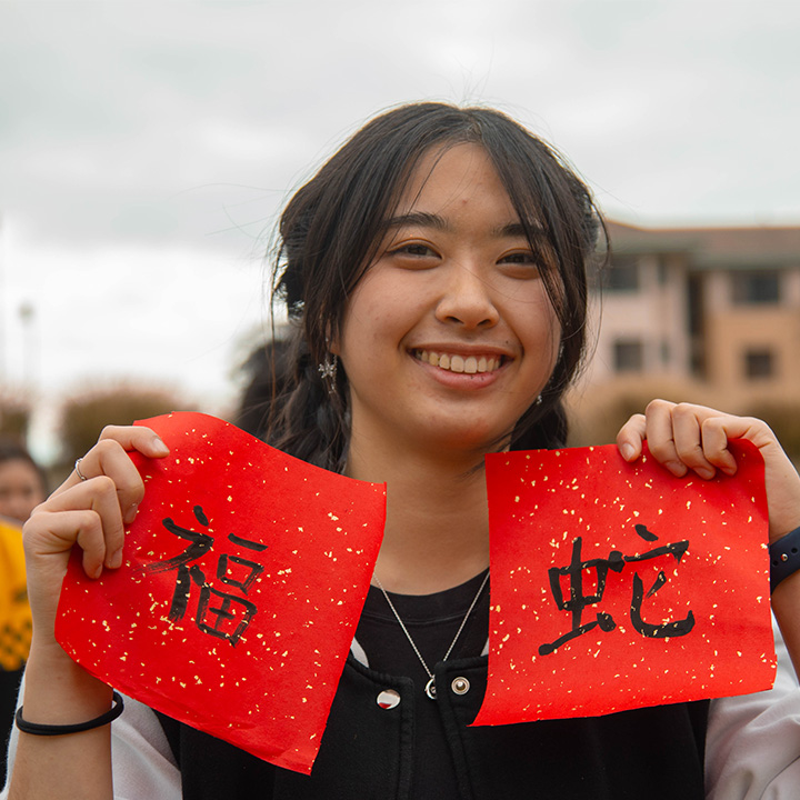 A student stands holds two red papers with hand painted characters on them.