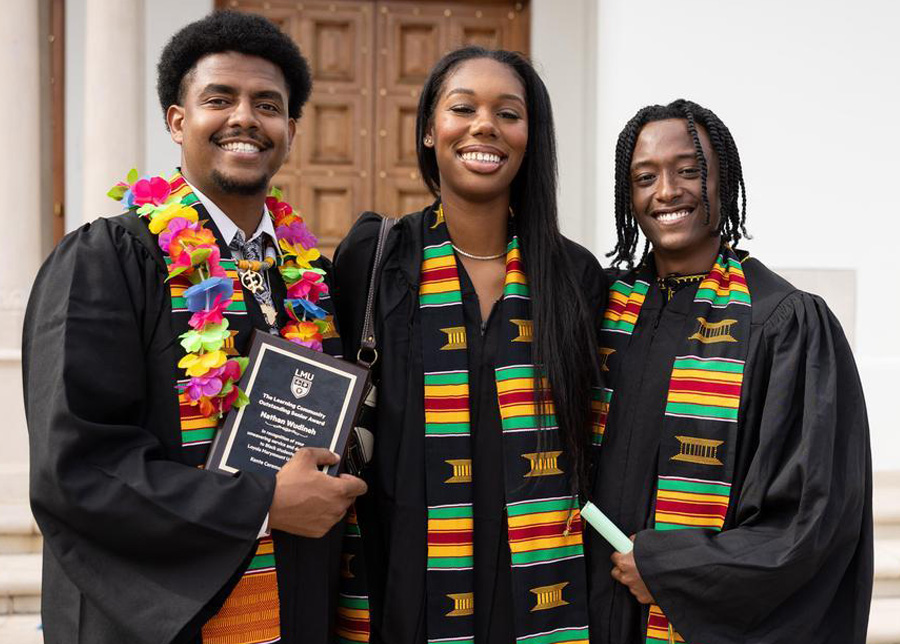 Three students pose together in grad regalia with their Kente stoles on in front of the chapel