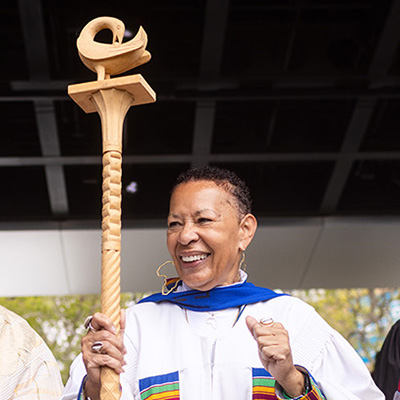 Cheryl Grills stands on stage in a white robe with a brown staff for LMU's annual Kente celebration.