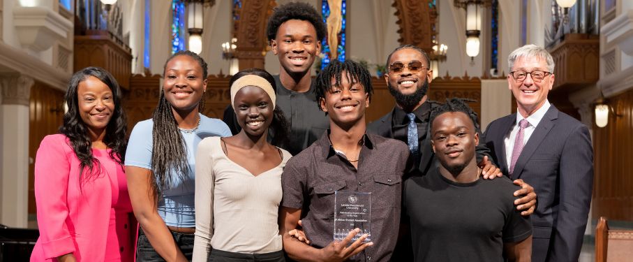 A small group of students stand on stage at the chapel with two professional staff to receive an award.