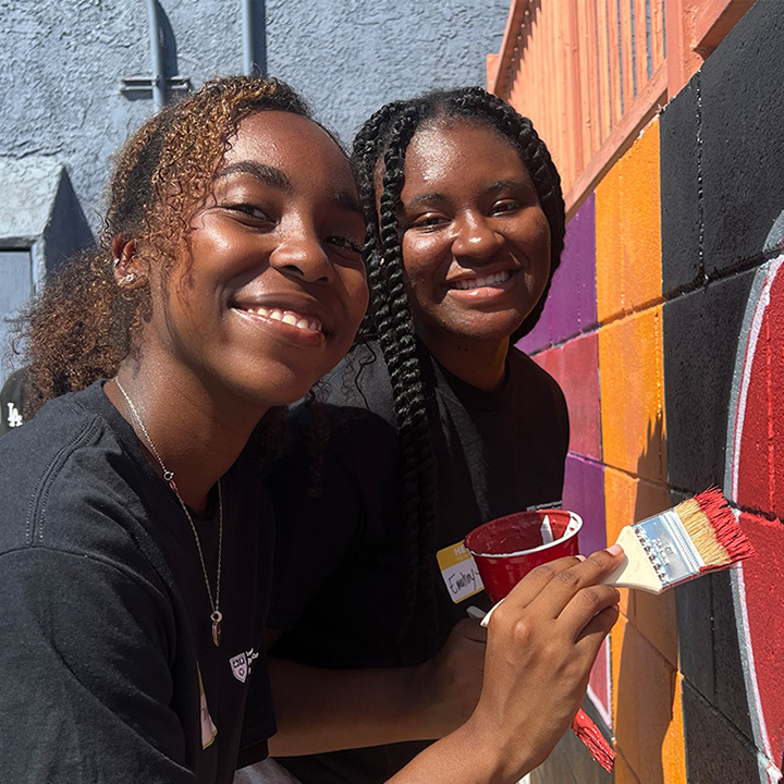 Two students in black t-shirts paint a colorful wall at a community service project.