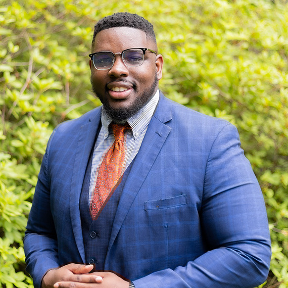 A headshot of Jason Perry in a blue suit in front of green bushes