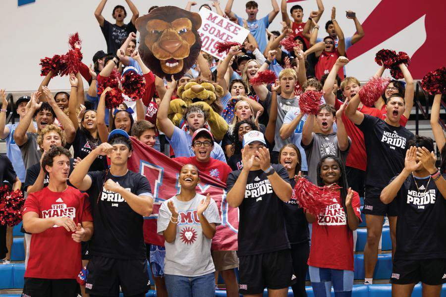 Fans stand cheering on the Lions during basketball wearing red, grey, and blue and holding up signs with Iggy the mascot's face on them.
