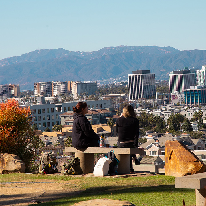 Two students sit on a bench near the bluff talking with green grass and orange landscape in the distance.
