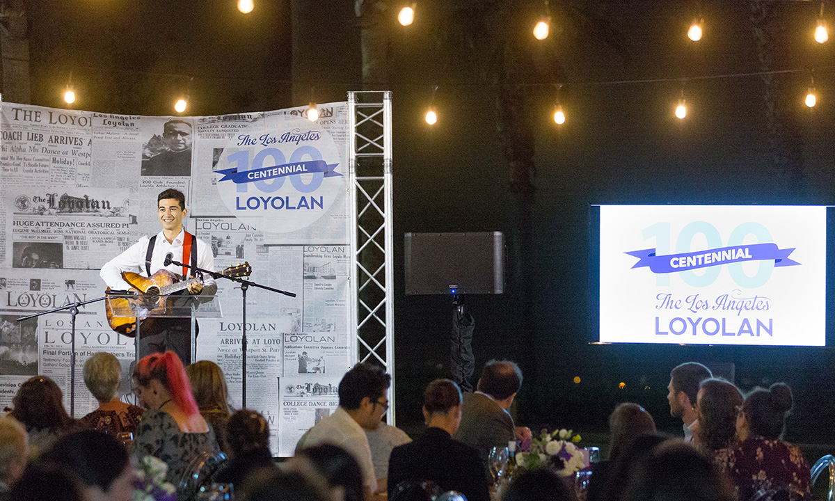 Students, staff, and guests gather outside at night to watch a student play guitar at the Loyolan Centennial celebration.