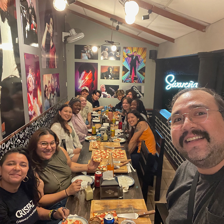 A group of students and staff gather at a table eating pizza.