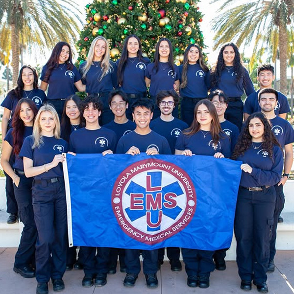 Student EMTs pose with a EMS blue flag in blue uniforms in front of the Christmas Tree on Regents Terrace.