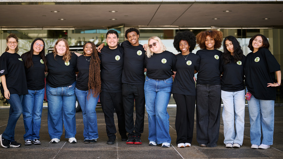 A group if students and two staff members stand outside of University Hall wearing black shirts and jeans.