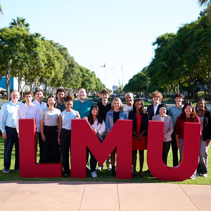 A group of students and staff pose in front of the LMU red letters outside on Regents Terrace.
