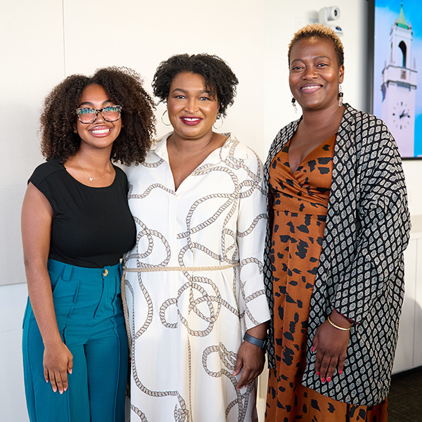 A student in a black shirt and teal skirt poses with Stacey Abrams in a white dress and a faculty member in an orange dress.