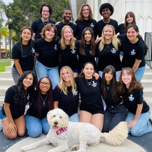 A group of students pose outside in black shirts with Buster, LMU's support dog.