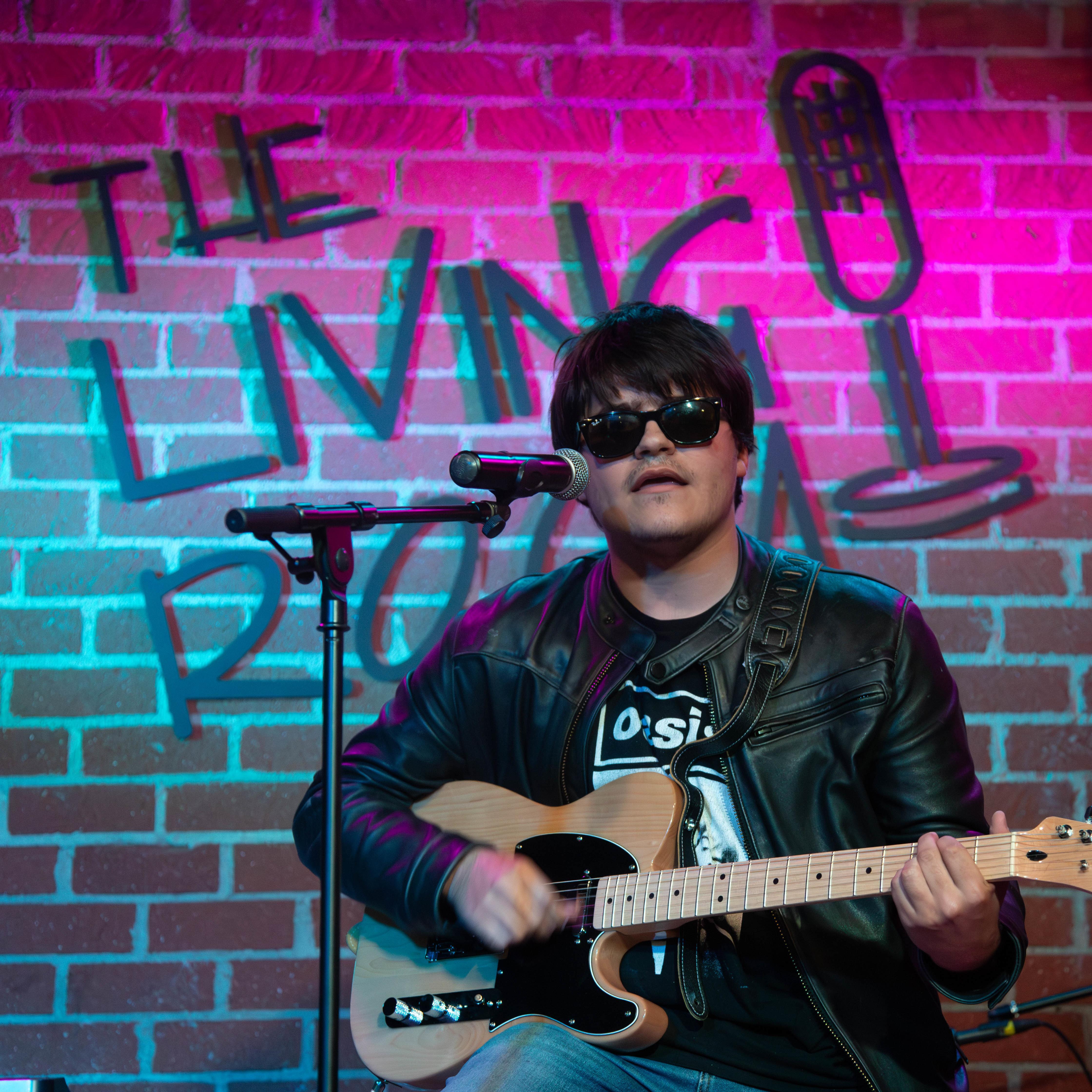 A male student in all black plays guitar on stage in The Living Room.