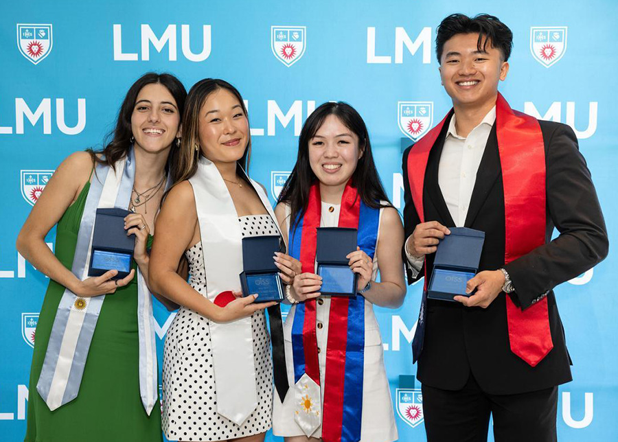 Four students pose in front a blue banner with LMU wearing international flag stoles during the International Grad Celebration.
