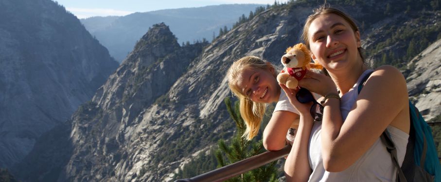 Two female students pose in front of a mountain holding little Lion stuffed animals.