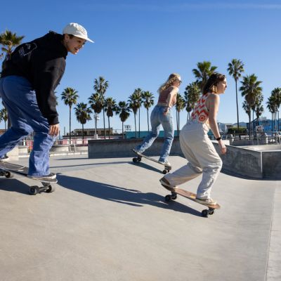 Three female students skateboard at the beach wearing colorful clothing during a sunny day with blue skies.