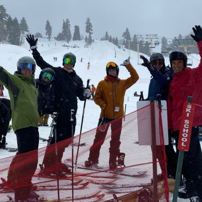 A group of students and staff pose on a mountain covered in snow with ski and snowboard gear on.