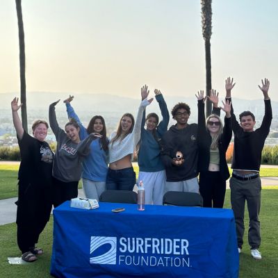 Students raise their hands standing outside in front of a blue tablecloth that says Surfrider Foundation.