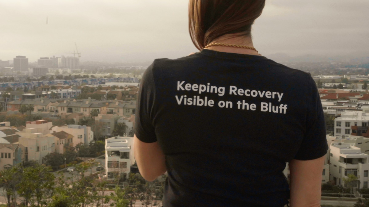 A person stands at sunset near the bluff with a black t-shirt on that says keeping recovery visible.