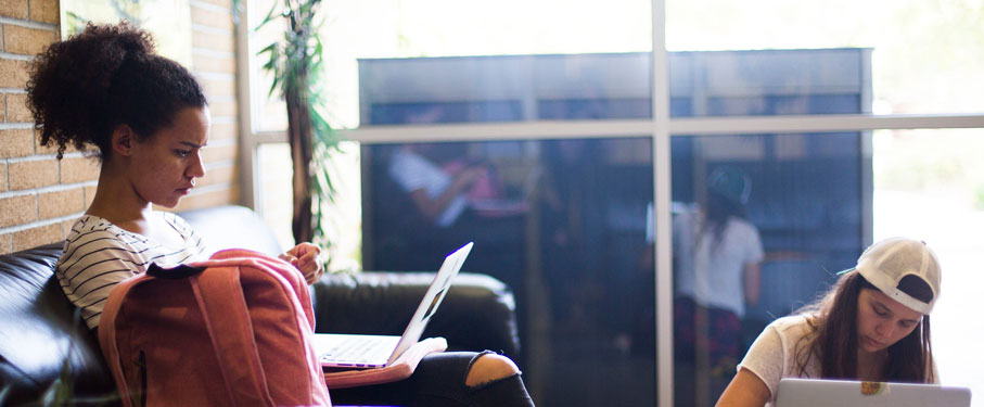 a student sitting on a sofa with laptop on lap and another student sitting below with laptop