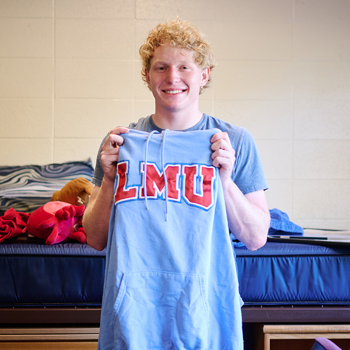 A student stands inside a dorm room holding a blue sweatshirt that had red letters for LMU