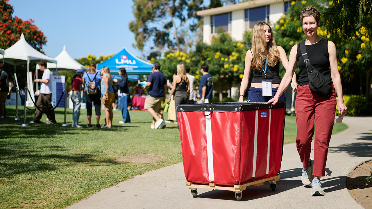 A person pushes a large red rolling cart filled with bags, boxes, and other move-in items along a campus walkway