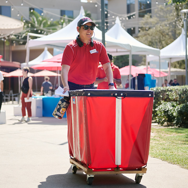 A person in a red polo shirt pushes a red moving cart across campus during move in.