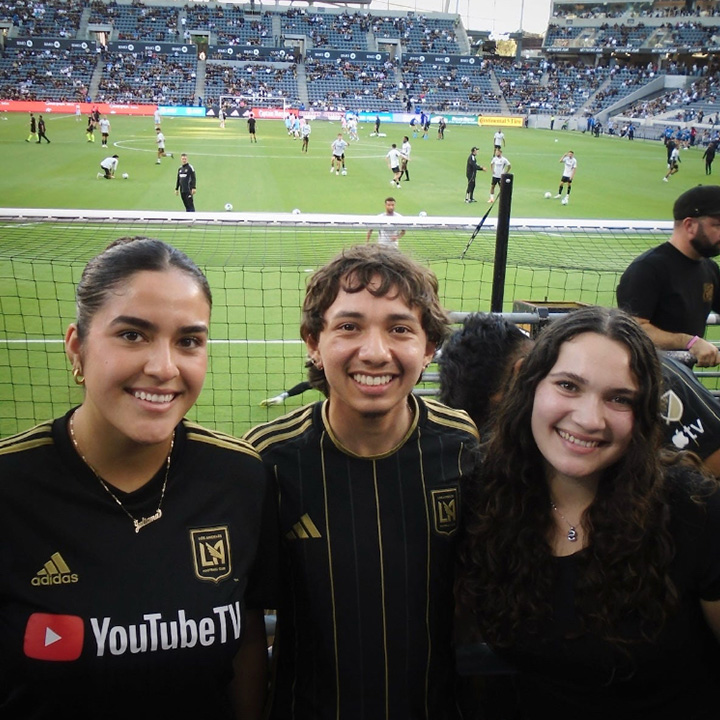 Three attendees posing in the front row of a soccer stadium wearing black LAFC jerseys
