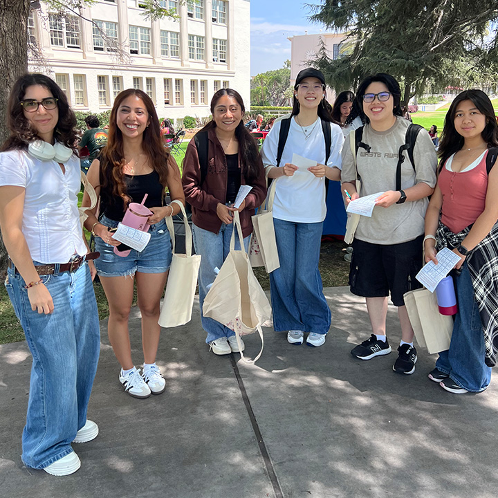 Six participants stand together on a shaded campus walkway during an outdoor event. Each person carries a canvas tote bag and holds a paper handout or card