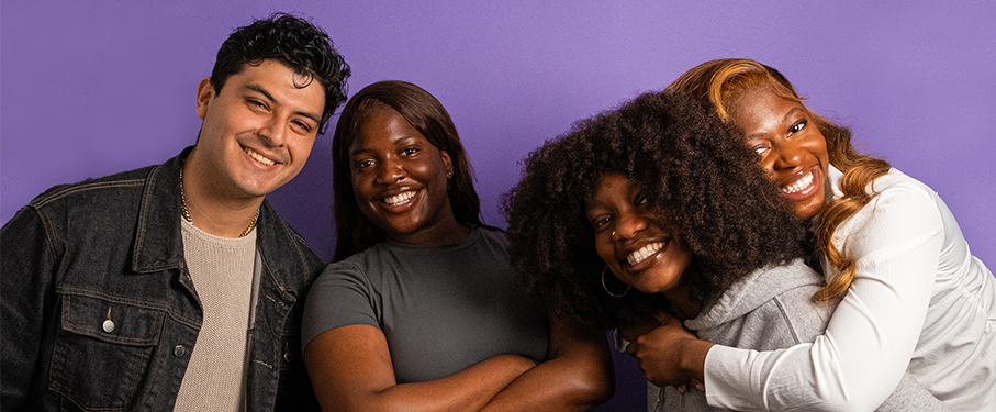 Four students stand in front of a purple background laughing and posing.