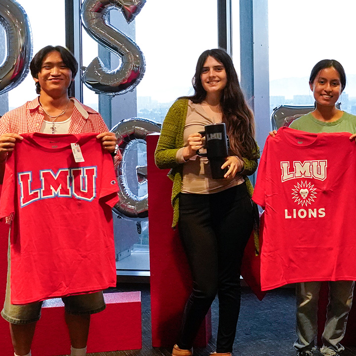 Three students stand in front of large silver balloon letters spelling LOS ANGELES and hold LMU-branded shirts and sweatshirts during a campus event