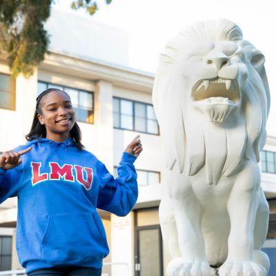 A student in a blue LMU sweatshirt stands next to the white Lion statue outside of Malone.