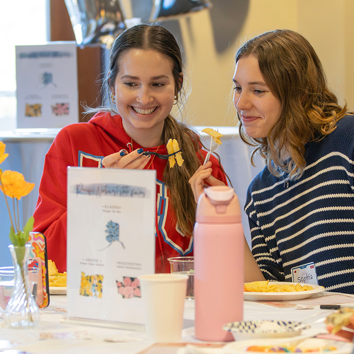 Two female students one with a red sweatshirt and one with a striped white and black sweatshirt pose with food on their forks.