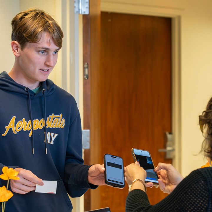 An attendee wearing a blue Aeropostale hoodie holds up a smartphone while standing near a registration or check-in table at an LMU Hometown Pride event